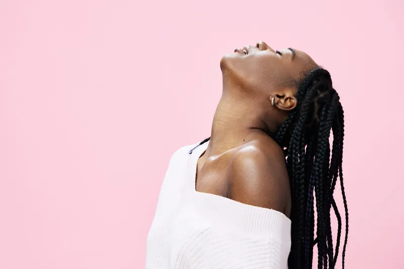 Young Black woman with braided hair, wearing a white sweater, exudes joy against a pastel pink background, perfect for fashion and emotional wellness themes