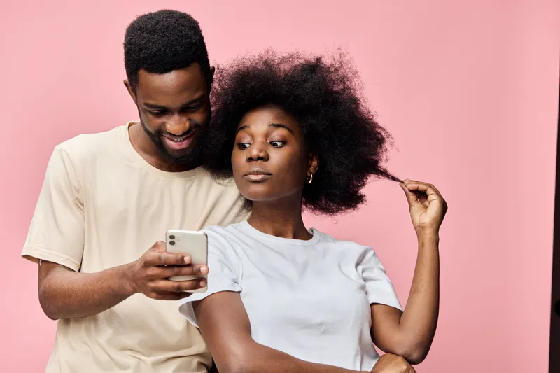 Young African couple joyfully interacting with a smartphone against a pink background, showcasing modern fashion and playful emotions, ideal for social media themes