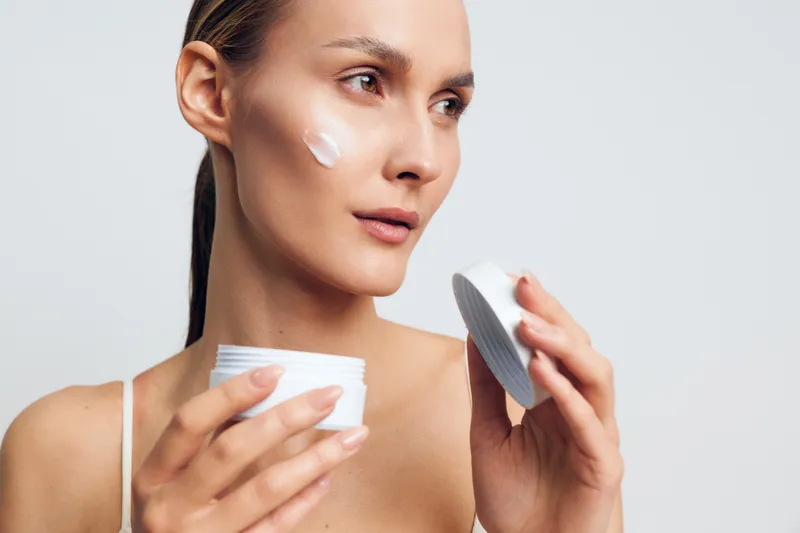 Young woman with clear skin applying white face cream on cheek. Skincare routine concept with natural beauty and self-care on a plain light background. Soft lighting highlights skin texture.