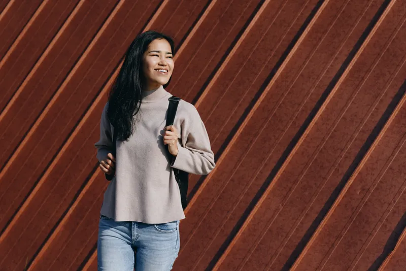 Woman with backpack in a casual sweater and jeans beside a diagonal wooden wall, cheerful expression, everyday exploration and urban chic style