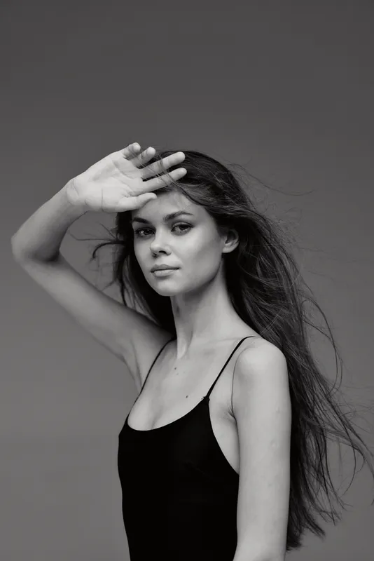 Black and white portrait of a confident woman with long hair shielding eyes in a minimalist studio setting.