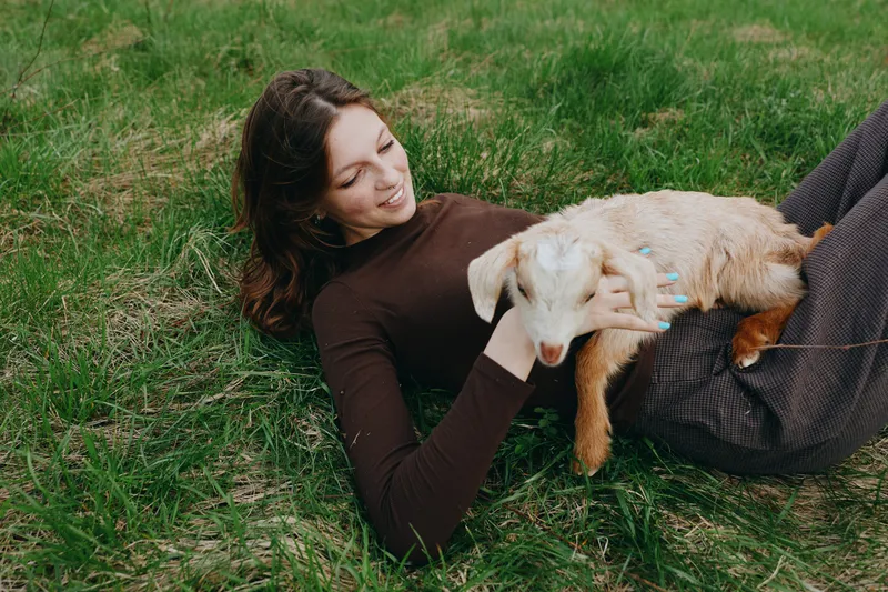 A woman in a brown sweater relaxes outdoors on grass, cradling a playful goat kid in her arms. A candid lifestyle moment highlighting affection and bond with animals.