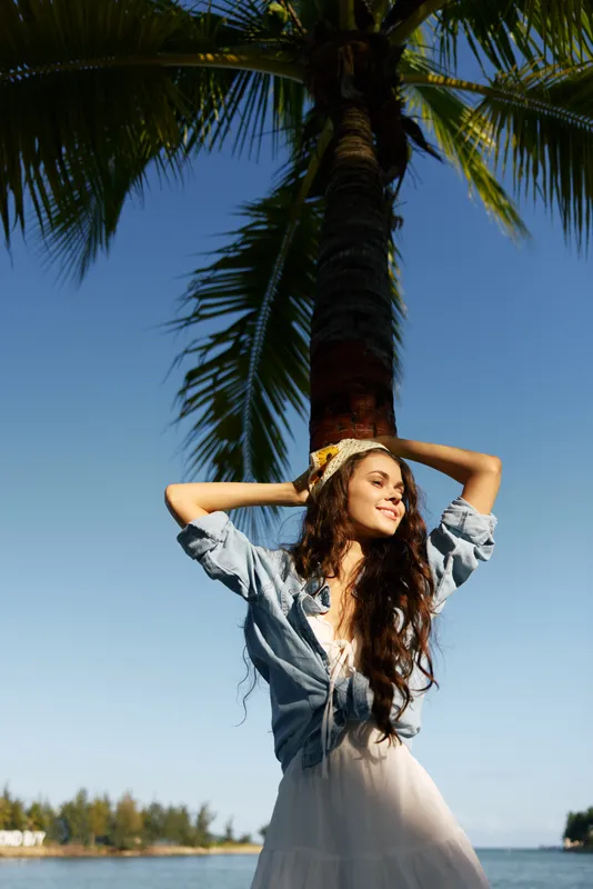 Young woman stands under a tall palm by the sea, wearing a light dress and denim shirt. Sunlit smile captures a carefree summer moment in tropical light.