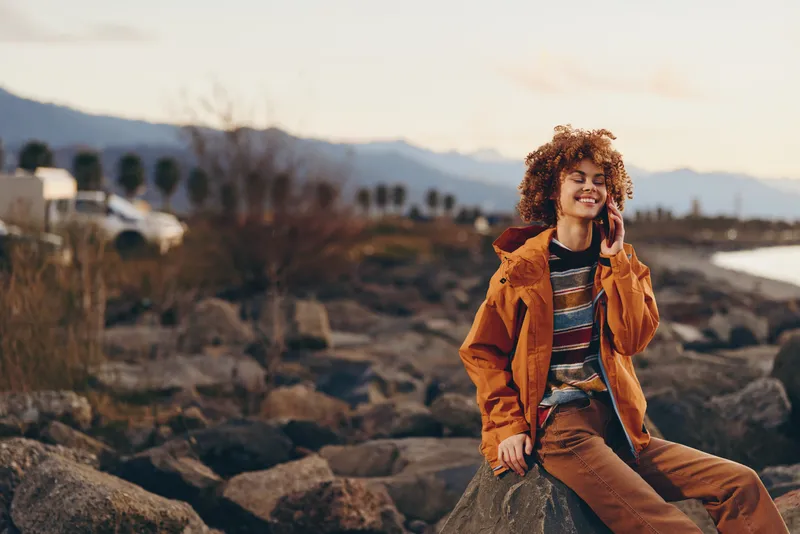 Smiling woman lifestyle outdoors on rocky coast at golden sunset, orange jacket, travel tourism mood, curly hair and relaxed hands on stones, mountains and soft sky in background