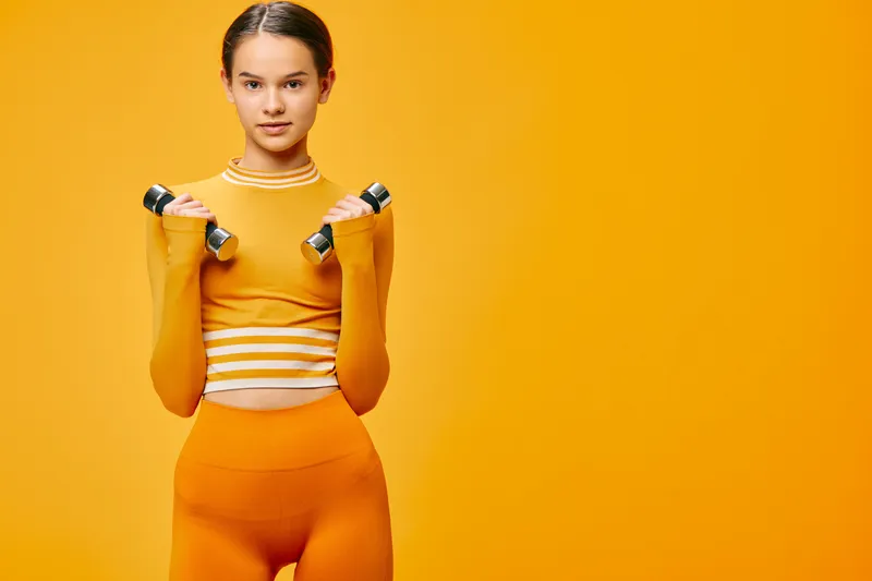 Female fitness model in orange athletic wear holding dumbbells for a workout, studio scene, strength training, active lifestyle, vibrant background, focused expression, full body pose, fitness