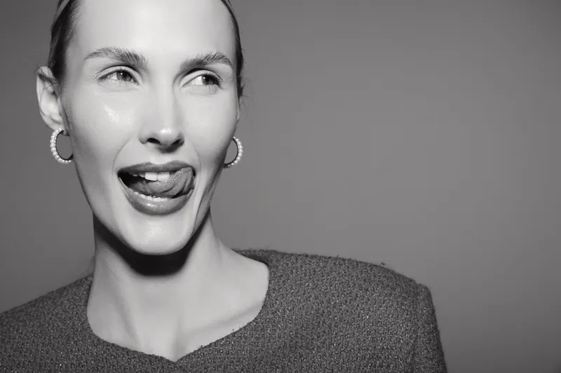Close-up of a smiling young white woman with fair skin and hoop earrings sticking out tongue, wearing a knitted sweater on plain background.