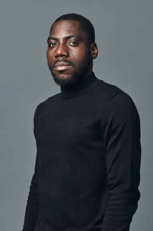 African American man with dark skin wearing black turtleneck sweater posing confidently against gray background in creative visual designer style portrait.