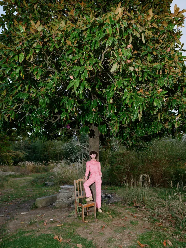 Young girl in pink overalls sits on a wooden chair beneath a lush tree in an open park, capturing a serene moment of solitude and nature, with soft light filtering through leaves.