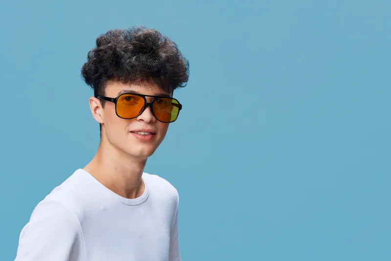 Smiling man wearing yellow sunglasses and white t-shirt stands against plain blue background with curly hair, casual style, youthful and cheerful appearance.