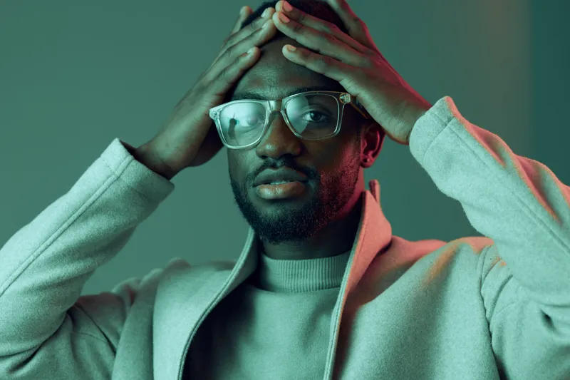 A thoughtful young man wearing glasses and a light sweater, expressing contemplation with a hand on his forehead against a soft green and pink background