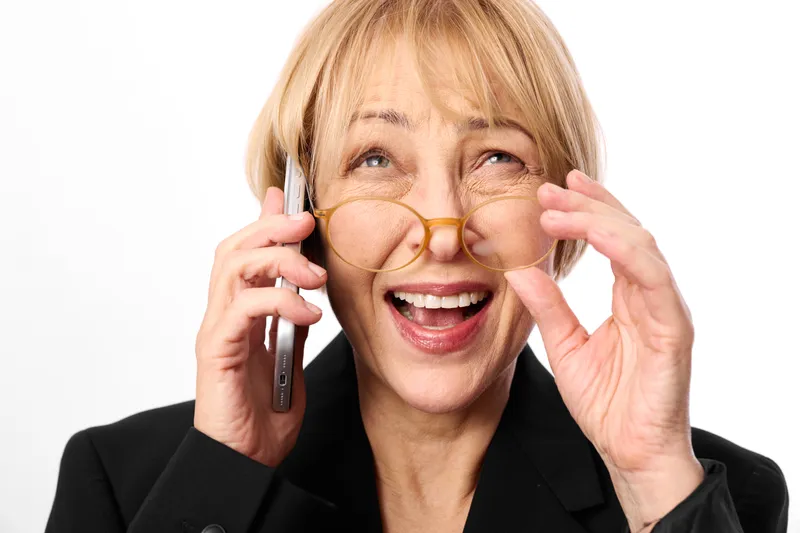 Cheerful older woman in business suit, laughing and speaking on phone, adjusting glasses, studio communication during a call, close portrait on bright white background