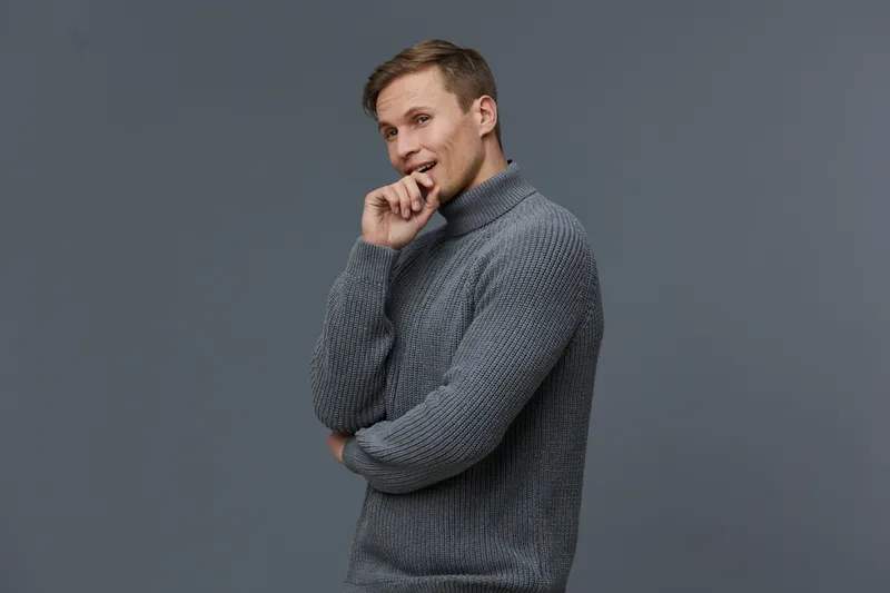 Smiling young man with casual gray sweater looking amused and thoughtful on plain gray background, posing confident and relaxed for a lifestyle portrait, indoor studio shot.