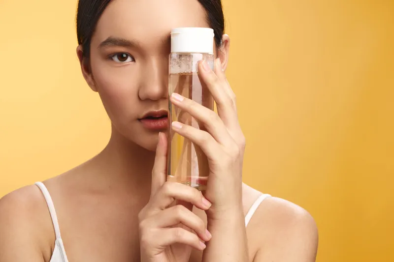 asian woman is shown holding a glass bottle of skincare product against a warm yellow backdrop, conveying a calm and focused beauty ritual in a modern setting.