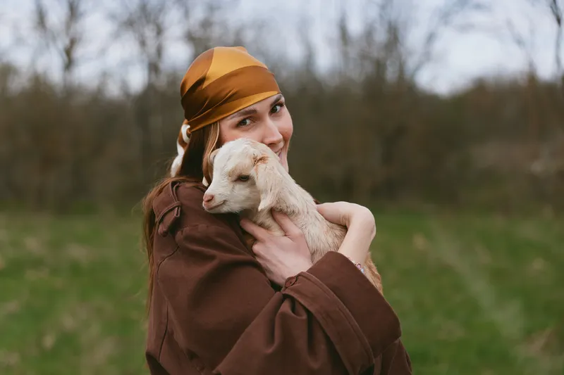 A woman in a brown coat and mustard headscarf embraces a young goat outdoors, capturing a warm countryside lifestyle moment with gentle affection and natural light.