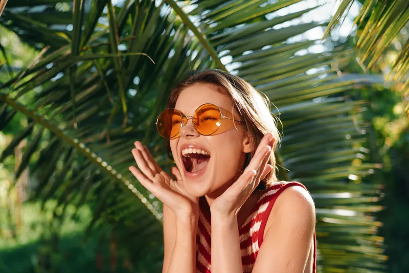 A cheerful young woman enjoys sunny outdoors, wearing sunglasses, with wide smile and open hands, expressing happiness and carefree energy in a vibrant leisure scene.