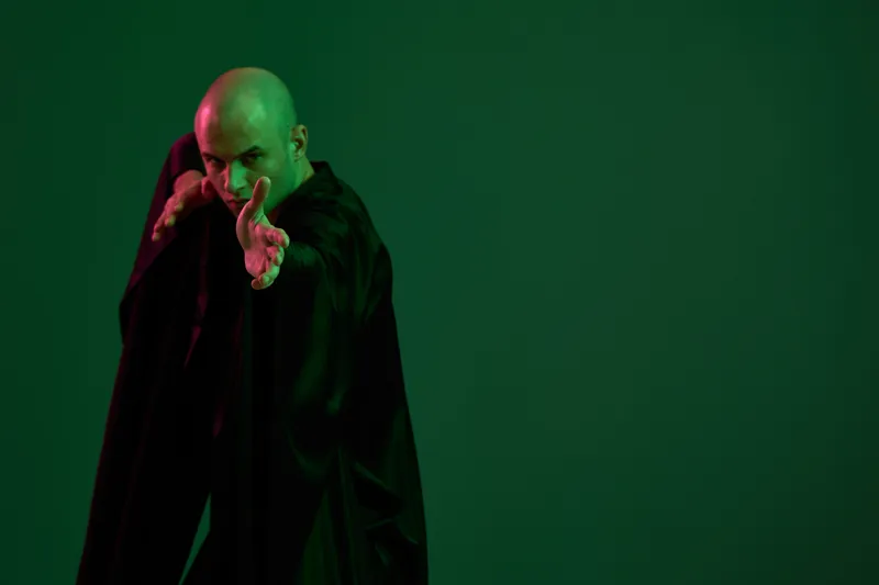 Mysterious man wearing a black cloak, posing dramatically against a vibrant green background, emphasizing intrigue and power in this striking composition.