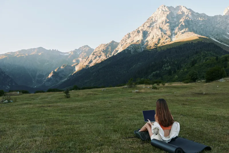 A person rests on a grassy meadow facing a majestic mountain range, reading a book and soaking in tranquil scenery, embodying focus, solitude, and outdoor inspiration.