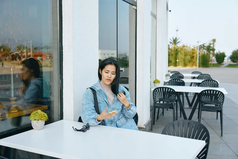 Woman at an outdoor cafe checks her phone beside a white table, sunglasses resting nearby, casual denim style, glass facade reflections, and empty chairs in a calm urban setting during daytime.