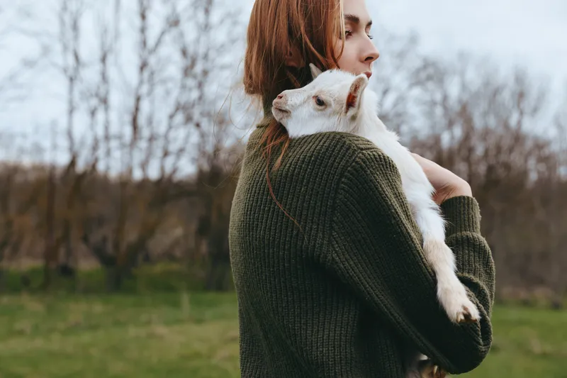 Young woman with long red hair holds a small white baby goat in her arms outdoors in a green sweater during an autumn day in nature.