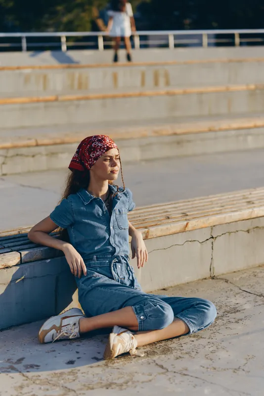 Lifestyle woman in modern boho-western denim street style sitting outdoors in film color, relaxed pose, warm sunlight, casual outfit with headscarf and sneakers.