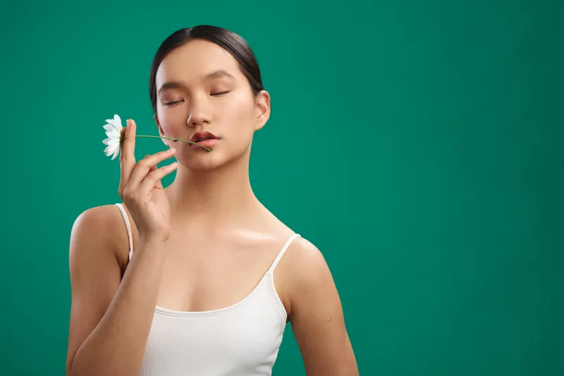 asian woman with a flower near her lips, eyes closed, posed in a simple studio against a teal backdrop, creating a calm, graceful portrait with soft light and subtle mood