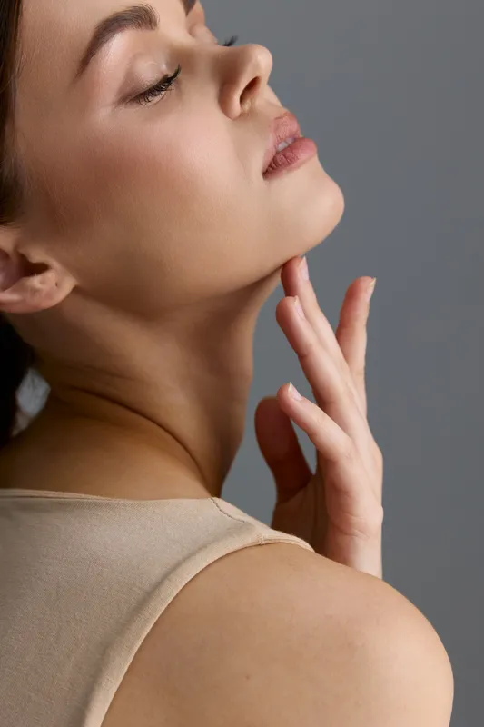 Beauty woman with smooth skin gently touches her chin, showcasing natural makeup and soft lighting on a minimalist neutral background, exuding calmness and elegance.