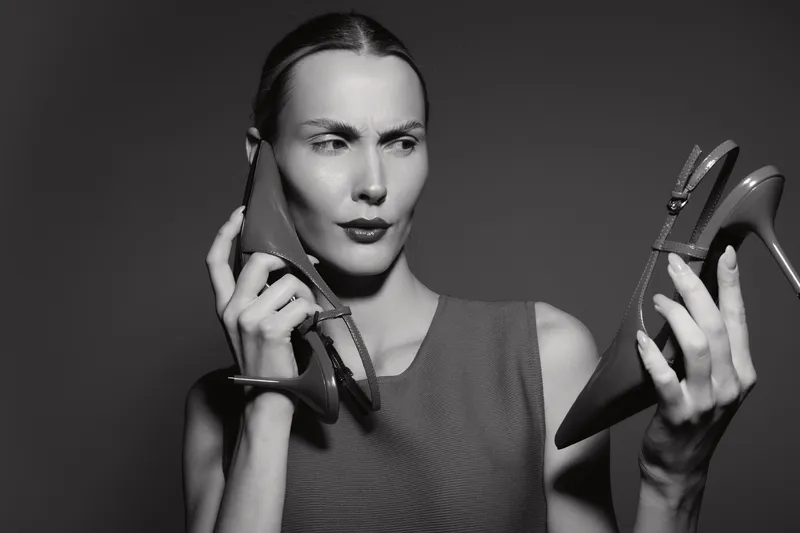 Caucasian woman with fair skin and dark hair holds two high heels, examining one closely with a thoughtful expression. Concept of fashion choice and style decision.