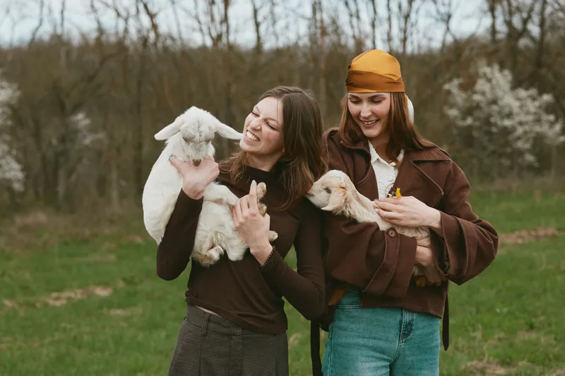 Two smiling women outdoors holding playful goats in a green field, showcasing casual countryside lifestyle, friendship, and animal companionship. Soft natural light creates a warm, wholesome mood.