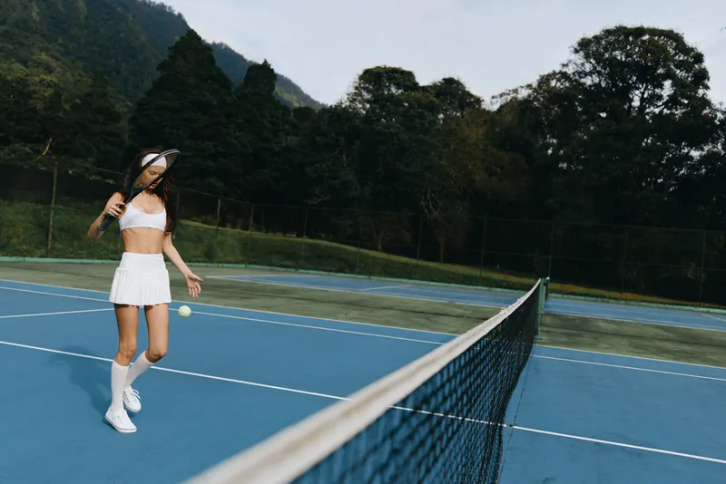 Young female tennis player in white sportswear plays on the blue court, showcasing her enthusiasm for the game amidst a lush green backdrop