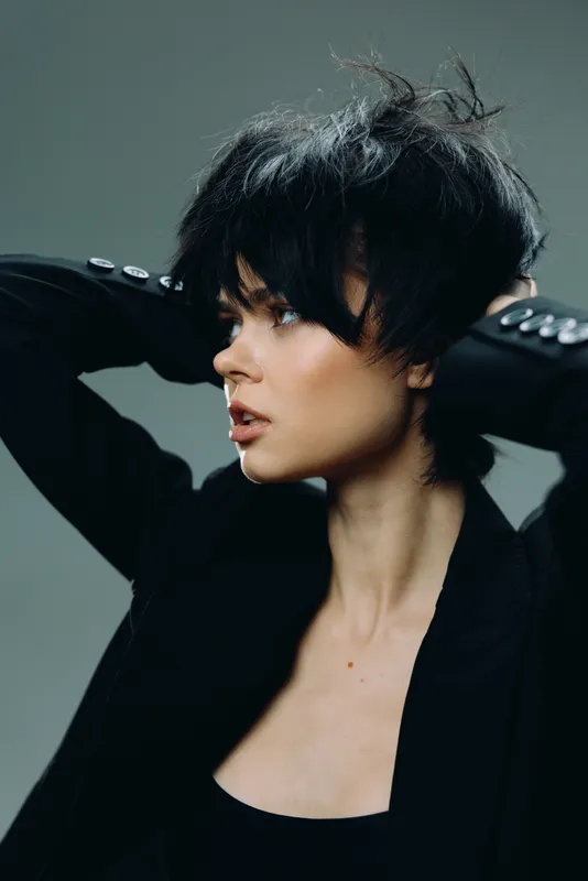 Stylish woman with short black hair in a black jacket posing against a dark background, showcasing modern fashion and confident expression.