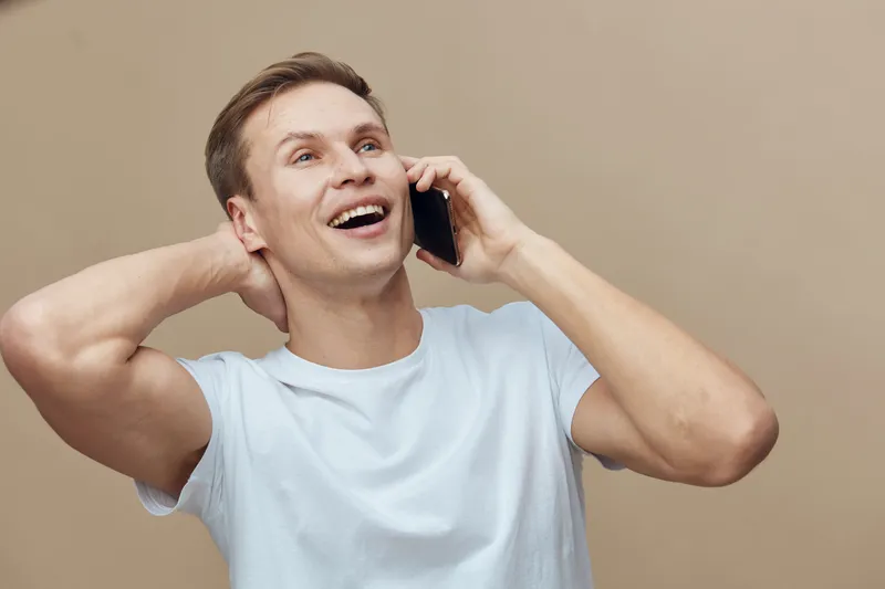 Smiling happy young man with short brown hair, wearing casual white t-shirt, talking on a mobile phone, cheerful expression, indoor neutral background, lifestyle portrait, male, relaxed mood, casual s