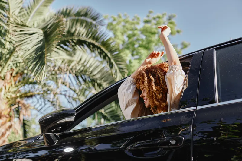 Smiling woman with curly hair enjoying a sunny day in a black car, surrounded by green palm trees and bright natural light outdoors.