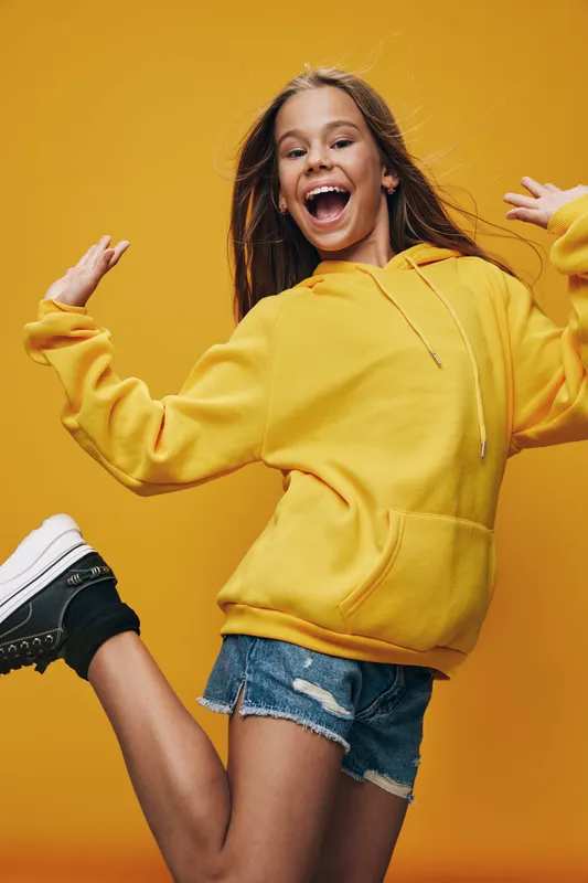 Girl in yellow hoodie and denim shorts jumping with joy on yellow background. Young female smiling, casual clothing and active pose with sneakers for lifestyle.