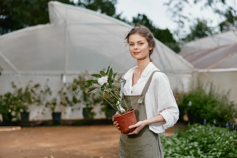 Young woman in overalls holding a potted plant in a greenhouse, showcasing sustainable gardening and a connection to nature