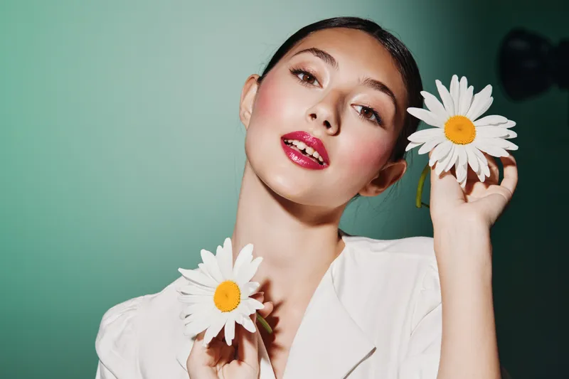 Creative beauty portrait of a young woman with bold red lipstick holding white daisies near her face. Soft studio lighting with pastel green sky background. Fashion concept.