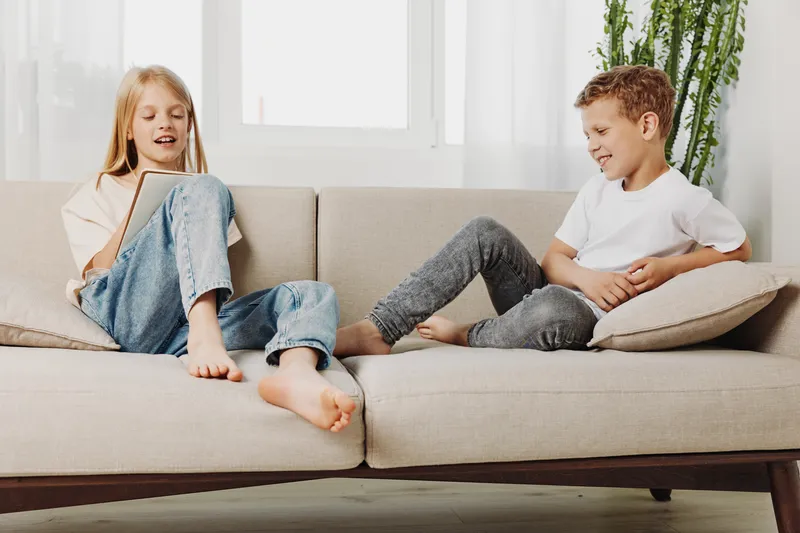 Two barefoot children sitting on beige sofa in bright living room, enjoying relaxed moment together. Casual clothes and natural light create warm, cozy family atmosphere with playful interaction.