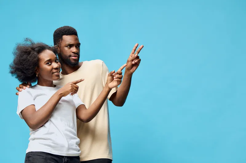 A young African couple joyfully posing against a vibrant blue background, showcasing playful emotions and casual attire in a modern lifestyle setting