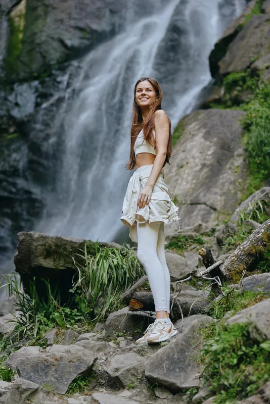 smiling woman enjoying nature at waterfall, wearing light sportswear and sneakers, surrounded by lush greenery and rocky terrain in outdoor adventure setting.