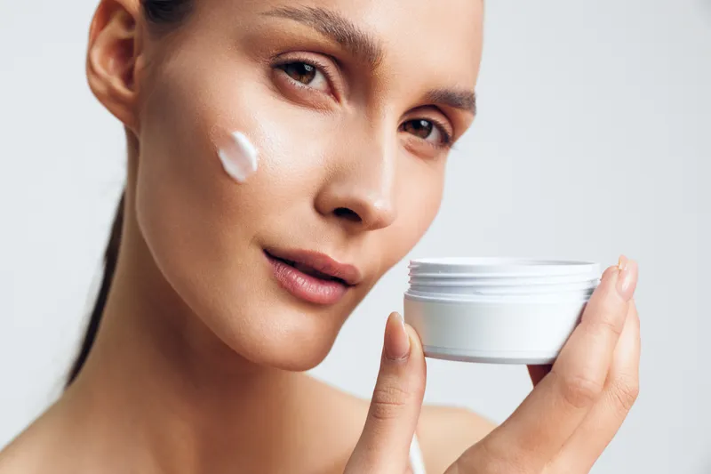 Young adult female with brunette hair applies cream to her cheek in a close up, conveying skincare, self care, and a calm soft lighting mood.