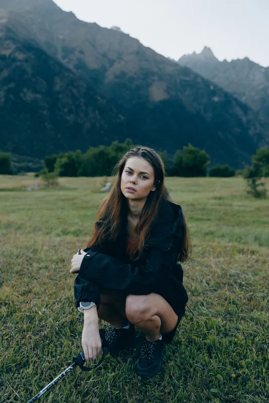 A solitary woman crouches in an open field with tall grass, wearing a dark jacket and boots, while distant mountains frame a calm, contemplative outdoor portrait.