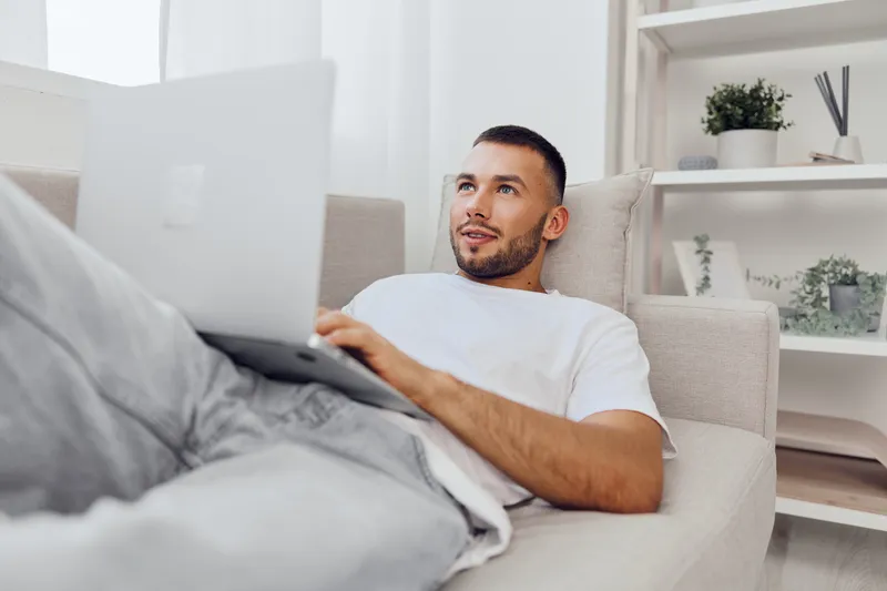 A relaxed man working on a laptop at home, embodying themes of solo living and emotional minimalism. The cozy atmosphere enhances productivity amid technology's demands.