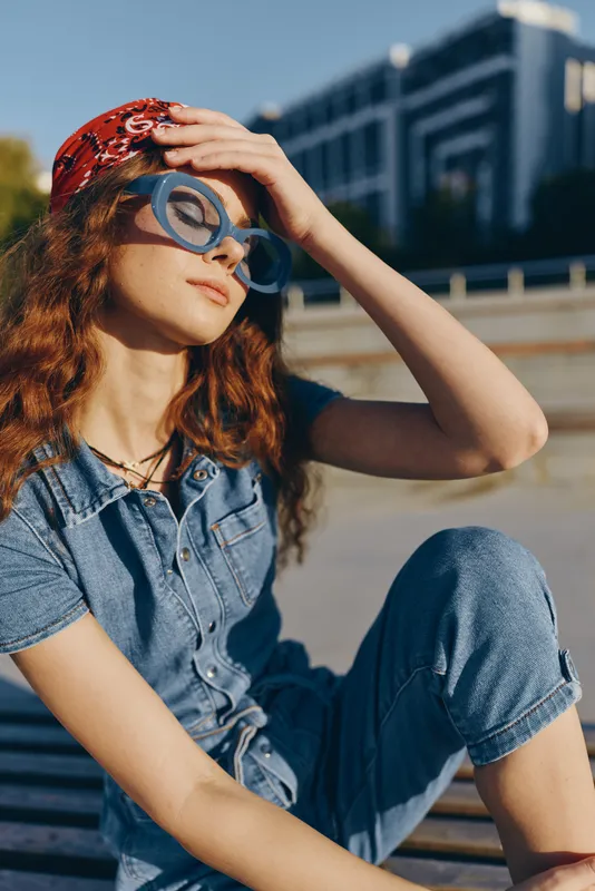 Lifestyle woman in modern boho-western denim street style wearing sunglasses and a bandana, enjoying sunny outdoor urban environment with film color tones.