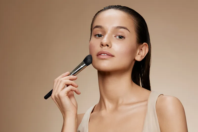 Young woman applying makeup with brush, natural beauty concept, neutral background, soft colors, enhancing self confidence, glow, skincare routine