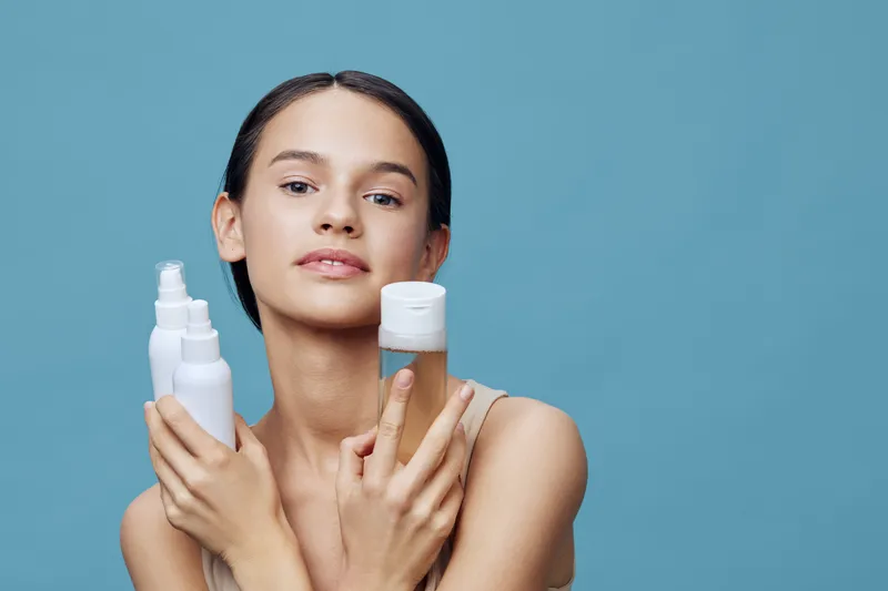 Young woman with radiant skin holding skincare products on a blue background, exuding confidence and beauty, ideal for wellness and skincare themes