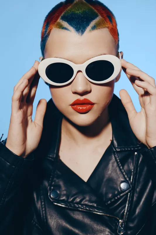 Trendy woman with colorful cropped hair wearing white retro sunglasses and black leather jacket, posing confidently against a bright blue background.
