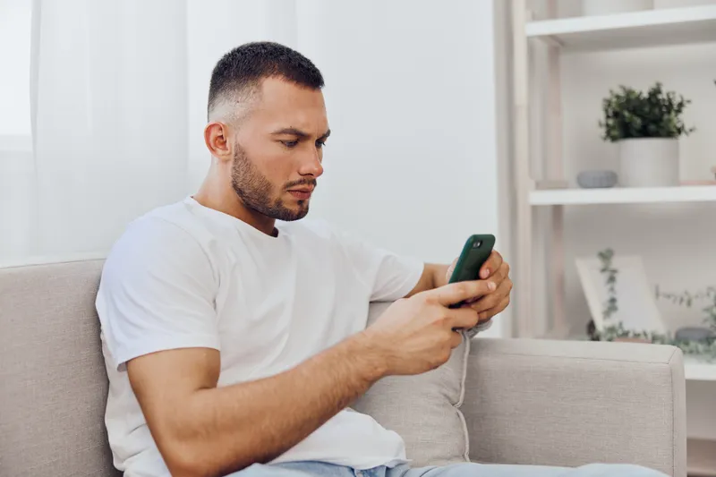Focused man using smartphone while sitting on a cozy sofa, bright interior, casual outfit, modern lifestyle, technology at home.