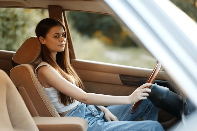 Young woman in casual outfit sitting in a vintage car, showcasing a pensive expression, surrounded by a natural backdrop, evoking a sense of freedom and adventure.
