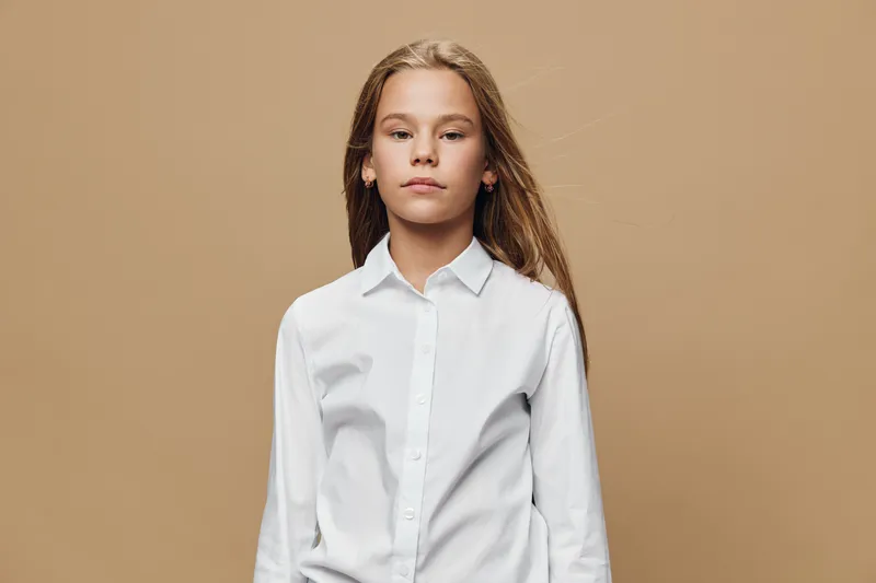 Portrait of confident girl with long hair wearing white shirt standing against beige background. Child with serious expression and casual hairstyle looking at camera indoors.