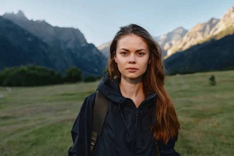 A focused young woman stands outdoors in a rugged landscape. She wears a dark jacket and backpack, looking toward the camera with calm confidence and quiet determination.