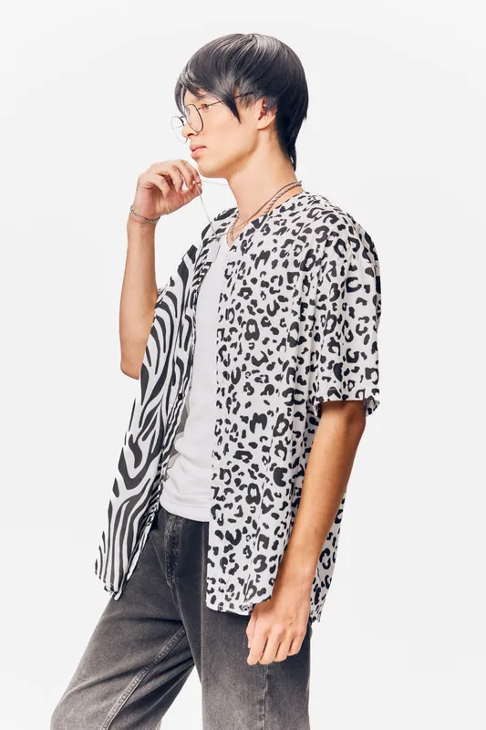 young man with glasses wearing a black and white leopard print shirt and gray jeans on a white background, thoughtful expression, casual style, isolated portrait
